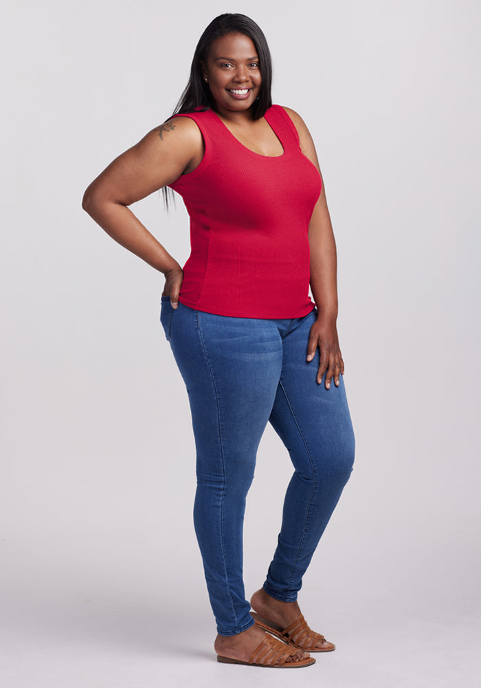 A woman with long dark hair, dressed in the Woolx Sloane Ribbed Tank in Poppy Spark, blue jeans, and brown sandals, stands smiling at the camera against a plain light background.