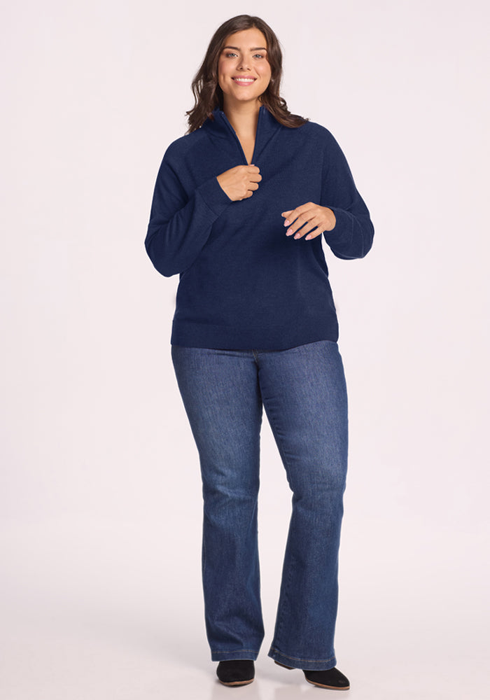 Woman smiles in a Woolx Collins Half Zip Sweater - Ocean Cavern, paired with blue jeans, against a white background.