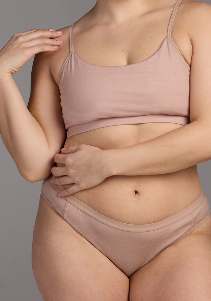 A woman models the Woolx Roxie Bikini Underwear in Morning Latte, standing against a gray background with her hands near her collarbone and abdomen to showcase its chafe-free Merino wool comfort. Her face is not visible.