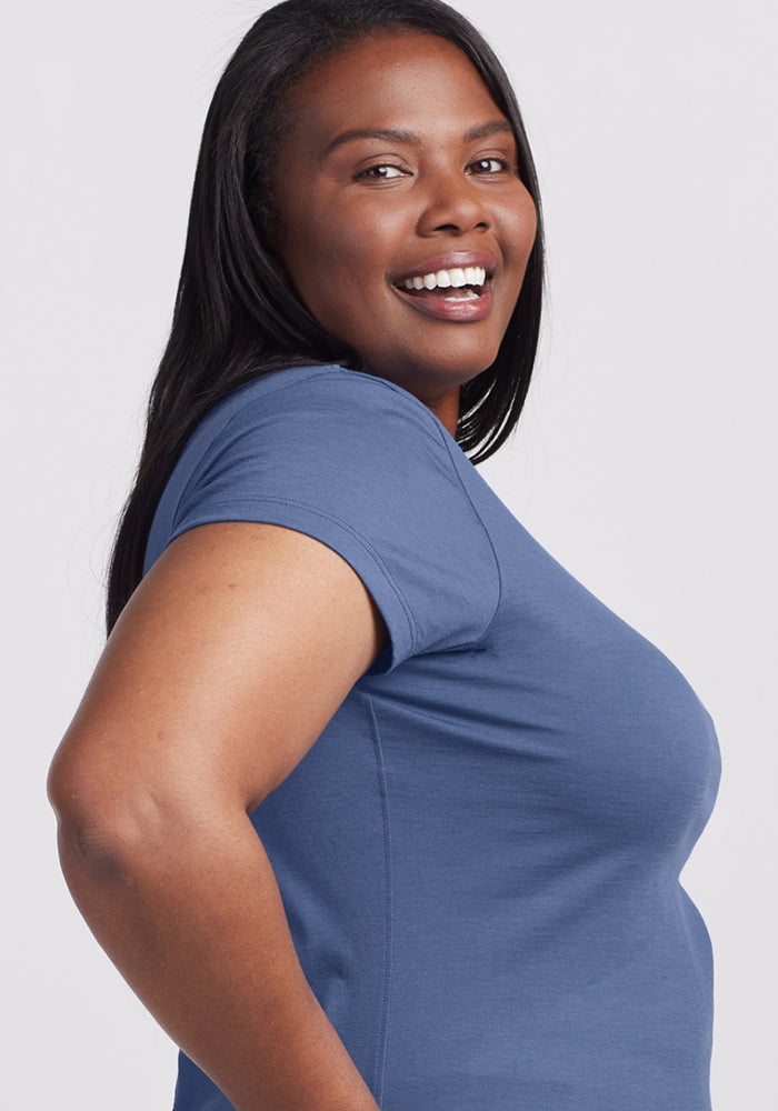 A woman with long dark hair smiles while standing sideways, wearing the Woolx Addie Crew Neck - Skyline short-sleeved merino shirt against a light gray background.