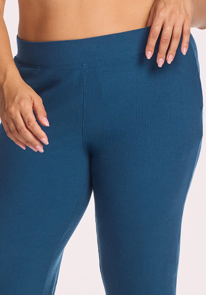 A close-up shows a person wearing Woolx’s Luca Ribbed Pants in Moroccan Blue, with one hand near the high waistband and neatly manicured nails, set against a plain light background.