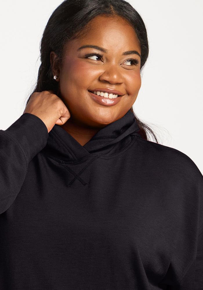 Woman smiles, looking aside in a Woolx Lizzy Hoodie - Black, featuring a buttery soft feel against a plain background.