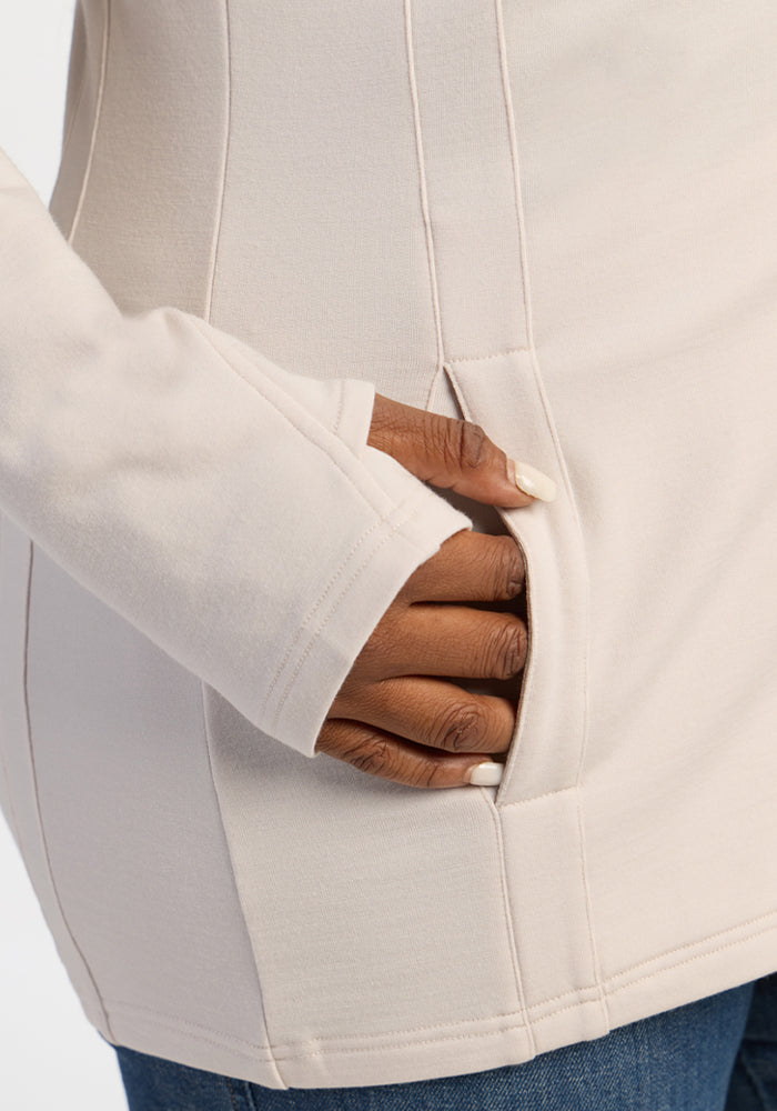 A hand with light-painted nails rests in the pocket of a Woolx Zoey Zip Up Hoodie - Ecru, paired with blue jeans.