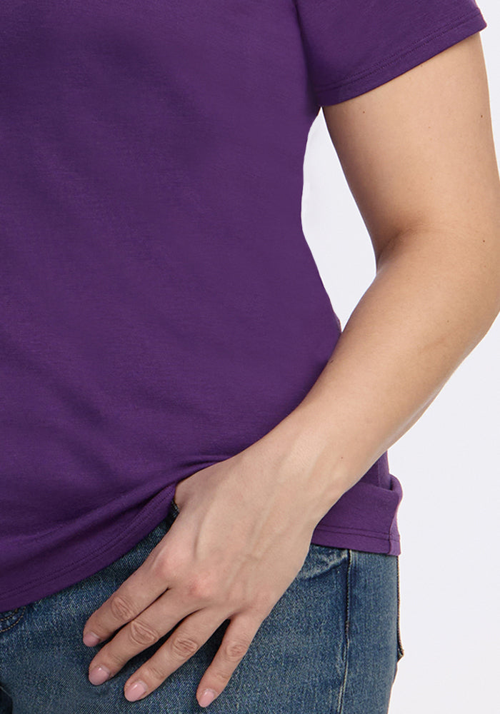 A person wears the Woolx Mia V Neck - Plum Crush t-shirt and blue jeans, resting their left hand on their hip against a plain light background.