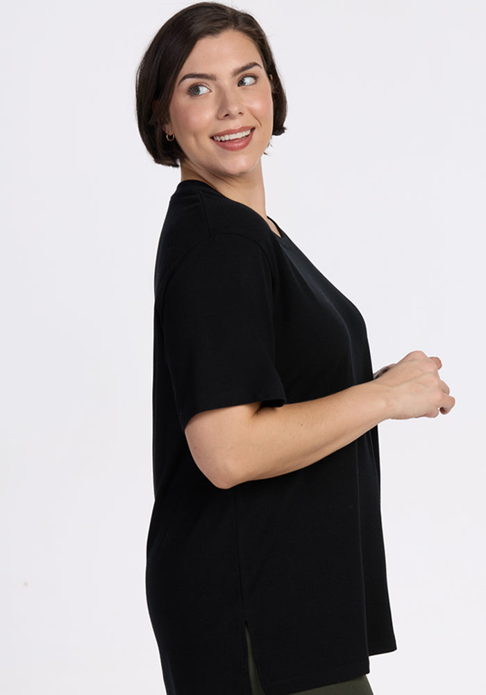A woman with short brown hair wears the Woolx Audrey Tee in black, standing sideways and smiling over her shoulder against a plain light background.