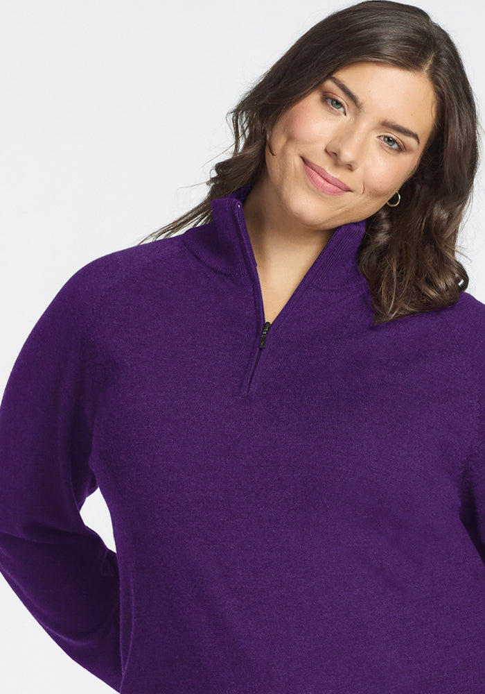 A woman with wavy brown hair smiles softly, tilting her head slightly to one side, while wearing the Woolx Collins Half Zip Sweater in Plum Crush against a plain light background.