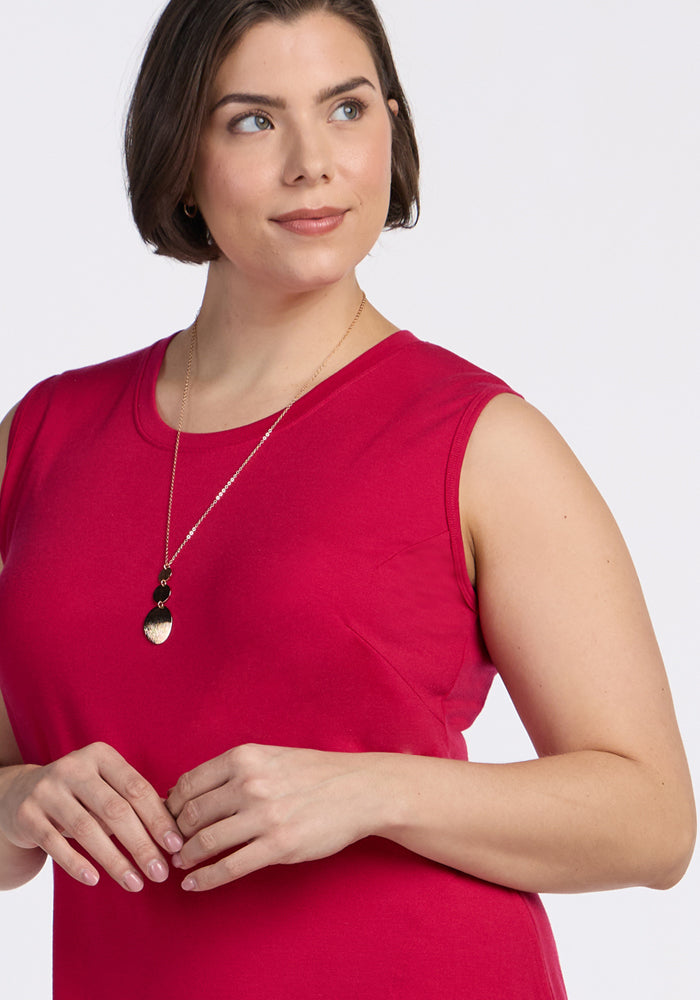 A woman with short brown hair models the Woolx Cassie Dress in Poppy Spark, a sleeveless bright pink travel dress, paired with a long gold necklace featuring black and gold pendants, as she smiles slightly against a plain white background.