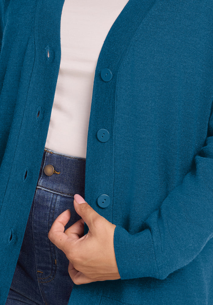 Close-up of a person in the Woolx Gia Sweater - Blue Pine over a white shirt and blue jeans, holding it near the buttons.