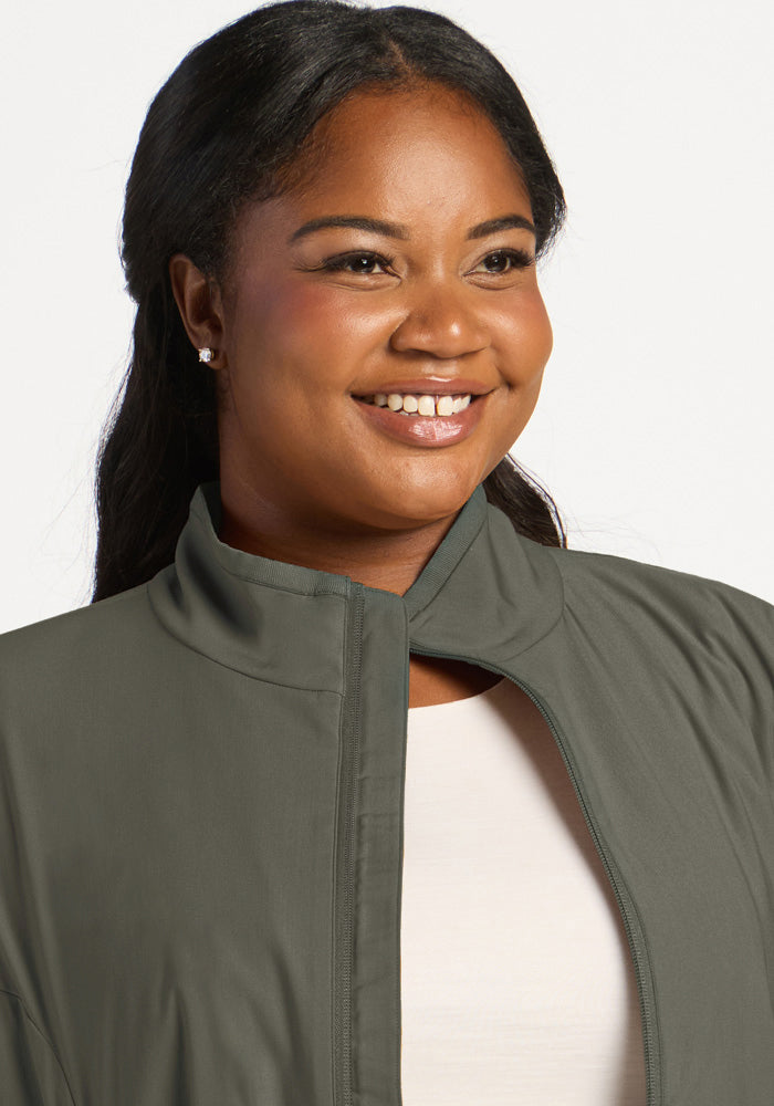 A smiling woman wearing a sage olive Woolx Cooper Windbreaker zip-up over a white shirt.