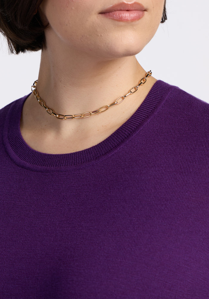 A person wearing a gold chain necklace and the Woolx Anna Crew Sweater in Plum Crush, with only the lower half of their face and neck visible.
