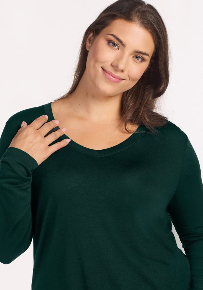 A woman wears a long-sleeved, dark green V-neck Lily V Neck Lounge Top - Deep Pine merino wool sleep shirt.