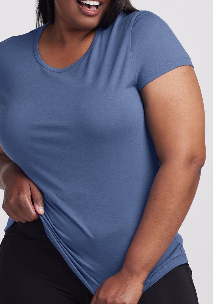 A woman smiles while holding the hem of her blue Addie Crew Neck - Skyline by Woolx, paired with black pants.