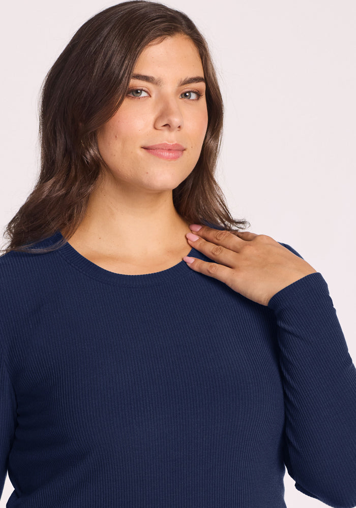 Woman with wavy brown hair in Woolx Hadley Ribbed Crew—Ocean Cavern smiles gently, hand on collarbone.
