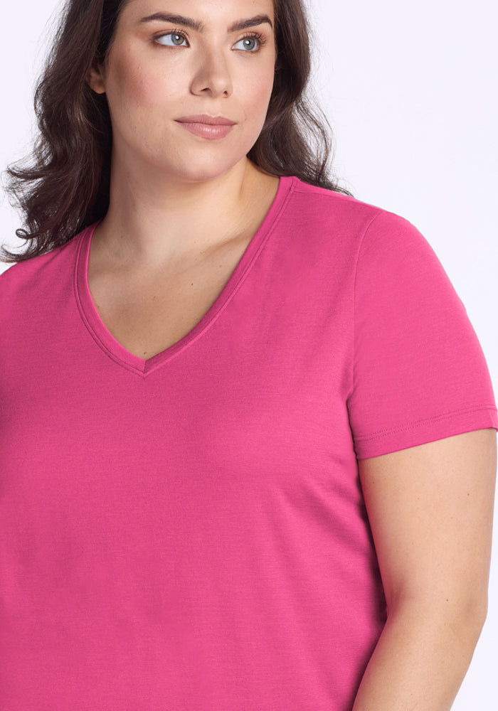 A woman with long brown hair models the Woolx Junie V Neck - Raspberry Fizz, a bright pink, temperature-regulating shirt with short sleeves, against a plain light background. She gazes slightly to the side.