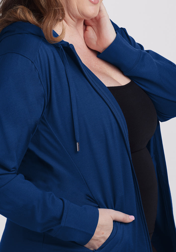 A woman models the Woolx Ryann Hoodie in Estate Blue over a black top, one hand in her pocket and the other touching her neck, shown from shoulders to mid-torso.
