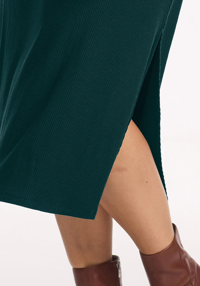 Close-up of a person in the Woolx Caroline Ribbed Twirl Dress in Deep Pine, paired with brown ankle boots.