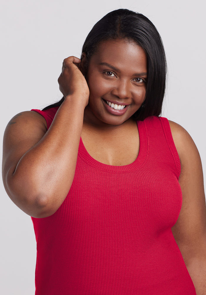 A woman in the Woolx Sloane Ribbed Tank in Poppy Spark smiles at the camera, touching her hair with one hand. She has long, straight hair and stands against a plain, light background.