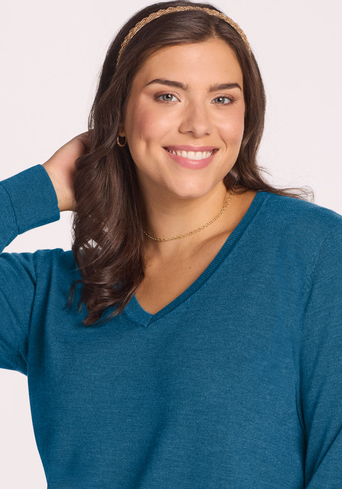 Smiling woman with wavy brown hair in a Woolx Francesca Sweater - Blue Pine, one hand behind her head.