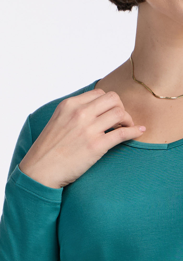 A person wearing the Woolx Layla V Neck Top - Surfside in teal touches their collarbone, showing a gold necklace. The plain background emphasizes the top’s moisture-wicking and odor-resistant qualities.