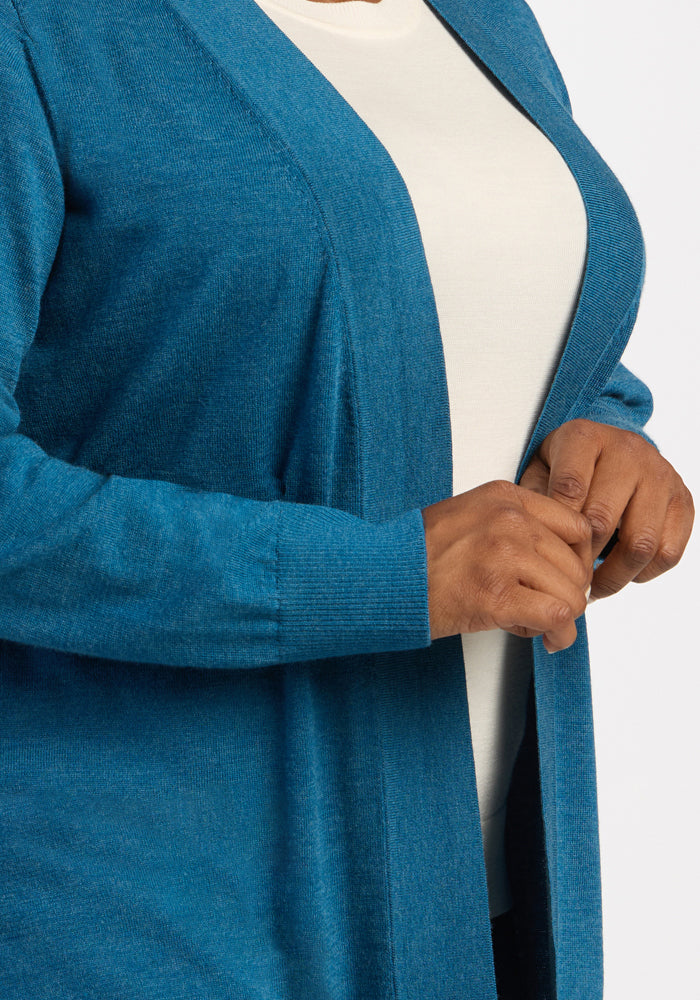 A person in a white shirt models the Woolx Macie Sweater - Blue Pine, holding its front edge with one hand.