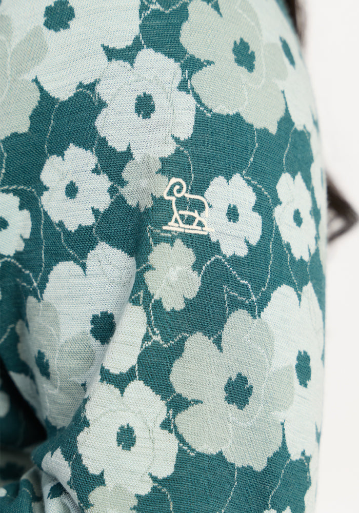 Woolx Emi 1/4 Zip - Coastal Wildflower: blue-green Merino base layer with floral design and white ram logo on shoulder.