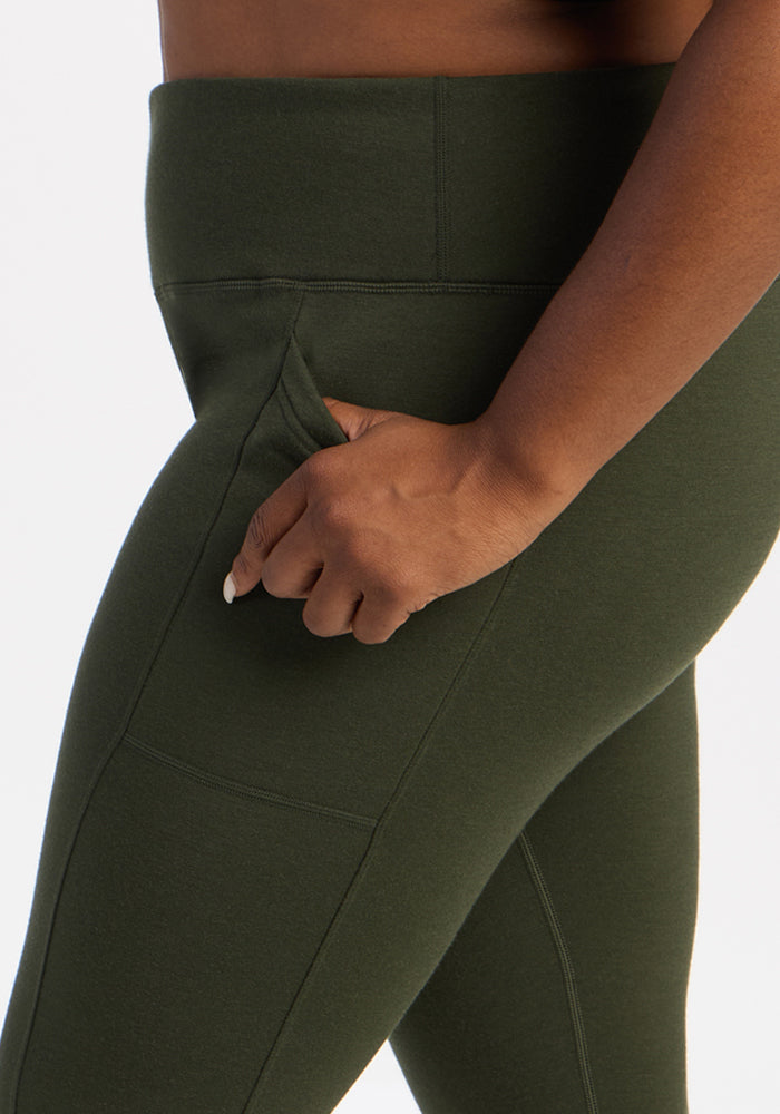 A hand slips into the side pocket of Woolx Frost Leggings - Duffel Bag, shown waist to thigh in olive green.