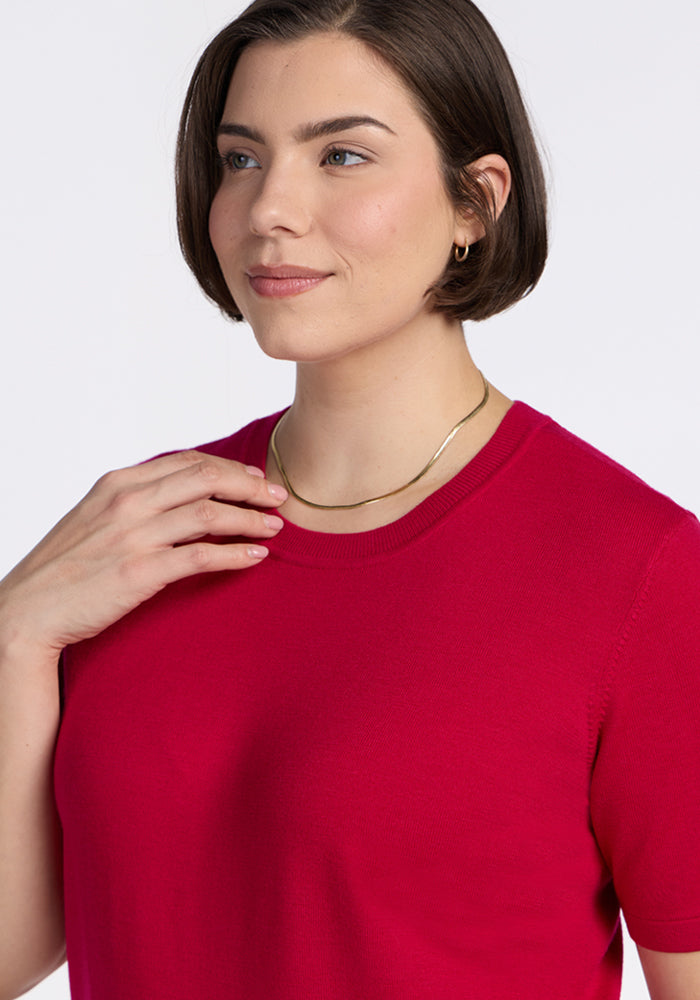 A woman with short brown hair wears a gold necklace and the Woolx Anna Crew Sweater in Poppy Spark, smiling softly with one hand touching her collarbone against a plain white background.