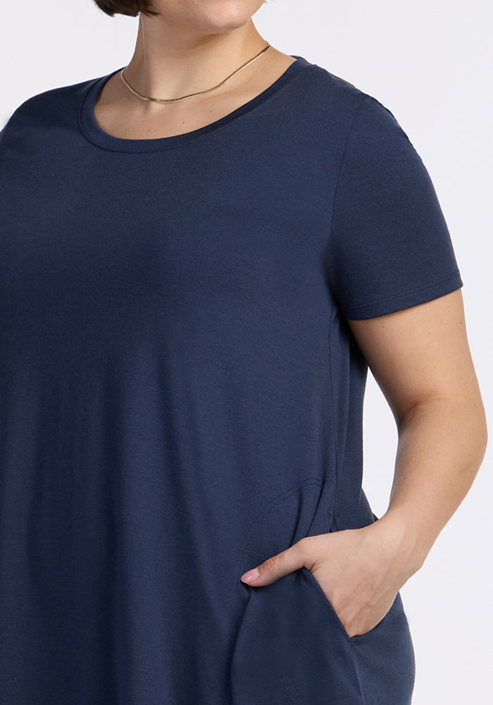 A woman wears the Woolx Georgie Dress in Deep Navy with short sleeves and a pocket, paired with a thin gold necklace. She has a short hairstyle and stands against a plain, light-colored background.