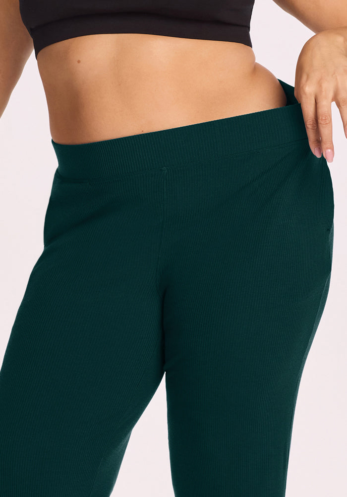 Model in a black top gently pulls the waistband of Woolx Luca Ribbed Pants - Deep Pine to show the comfy wide-leg fit.