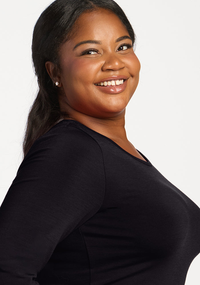 A woman with long dark hair smiles, wearing the Woolx Margot Bodysuit in black against a white background.