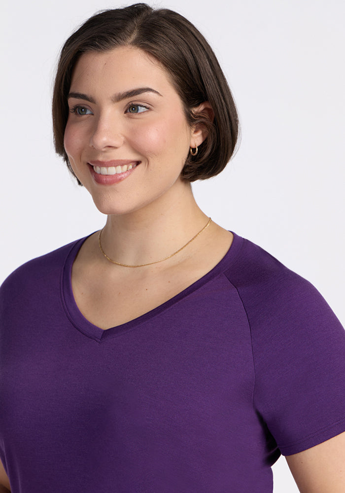 A woman with short brown hair, wearing the Woolx Mia V Neck in Plum Crush and a thin gold necklace, is smiling against a plain light background.