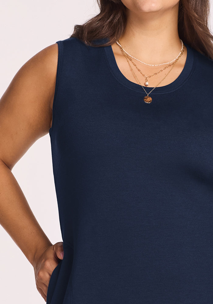 A woman models the Woolx Cassie Dress in Deep Navy, a sleeveless travel style with wrinkle-resistant fabric. She stands with one hand on her hip, layered gold necklaces visible as her face is partly out of frame.
