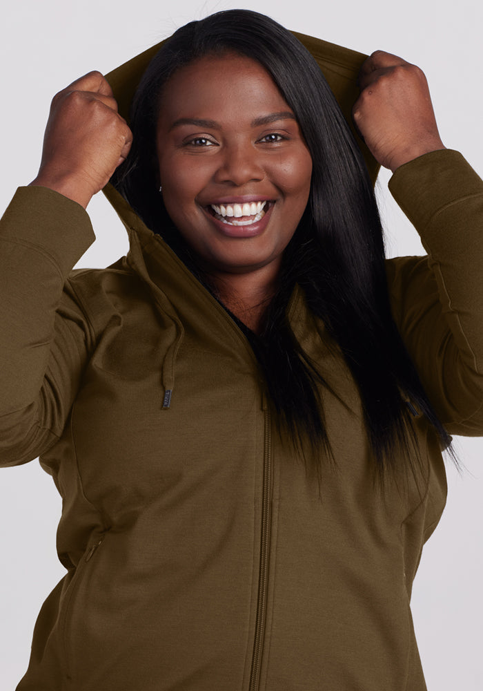 Smiling woman wears the Woolx Ryann Hoodie in Olive Leaf, pulling up the hood with both hands against a light background.