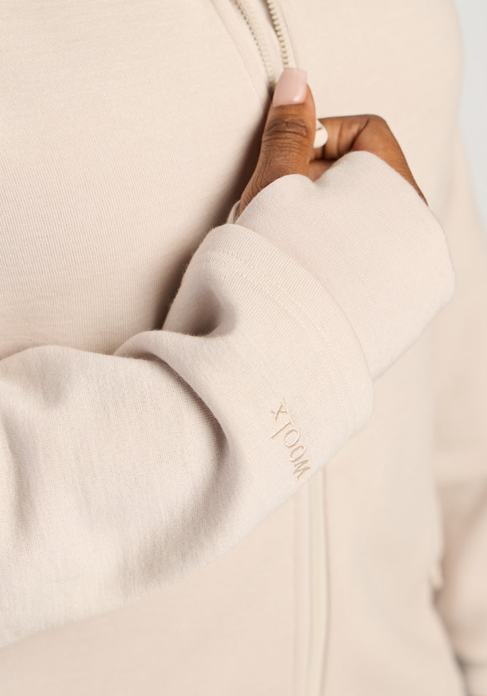 Close-up of a hand adjusting the cuff of an ecru Woolx Cubby Hooded Sweatshirt, showing an embroidered logo.