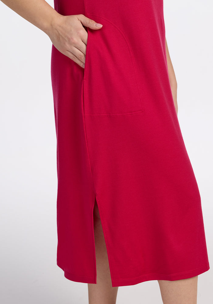 Wearing the Woolx Cassie Dress in Poppy Spark, a person slips one hand into a roomy side pocket. The bright, breathable dress has a smooth texture and a small hem slit, with just the lower torso and part of an arm visible.