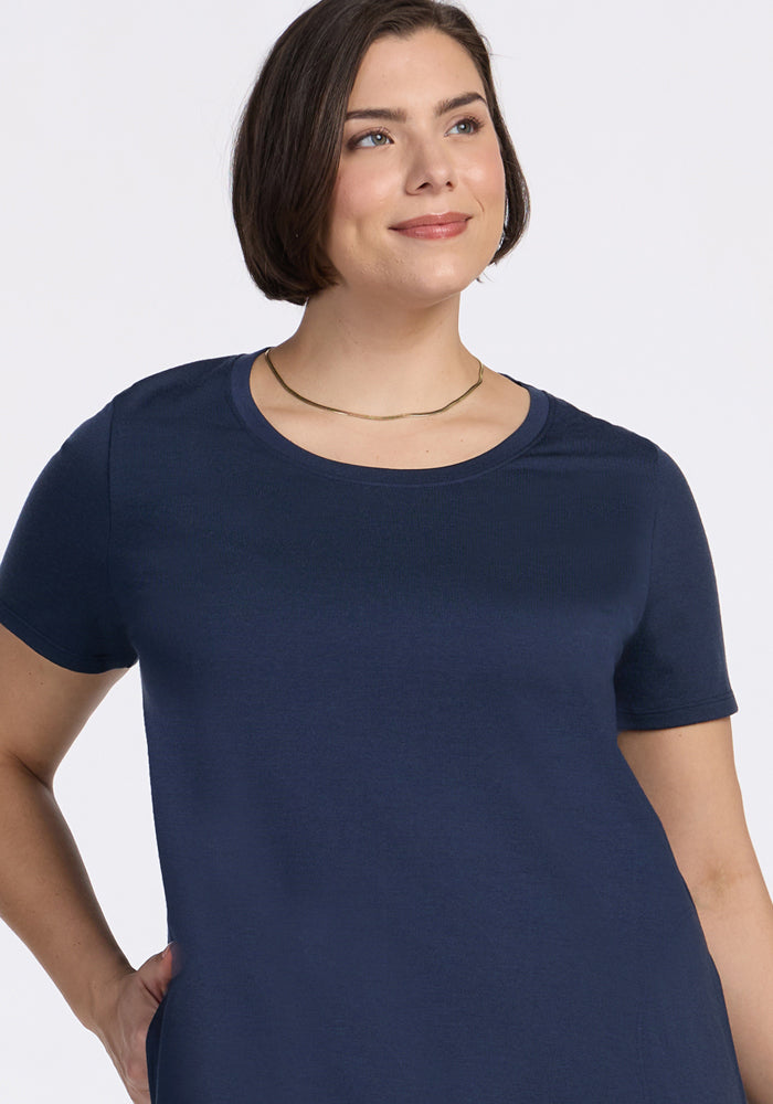 A woman with short brown hair, wearing the Woolx Georgie Dress in Deep Navy and a simple gold necklace, smiles slightly while looking up. She stands with one hand on her hip against a plain light background.
