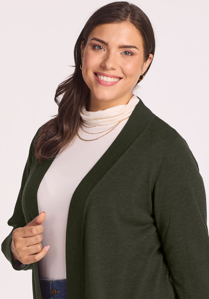 Smiling woman with long brown hair wears the Woolx Macie Sweater in Duffel Bag over a white top, plain backdrop.