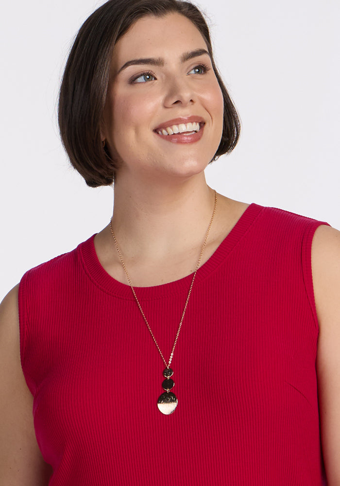 A woman with short brown hair smiles and looks to the side. She is wearing the Woolx Iris Ribbed Dress in Poppy Spark and a long gold necklace with black and gold geometric pendants.