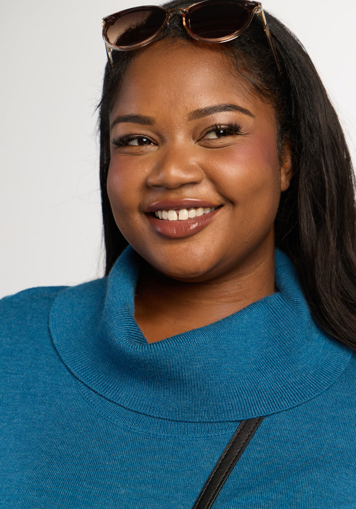 Smiling woman in Woolx Josie Cowl Neck Sweater - Blue Pine, sunglasses on head, neutral background.