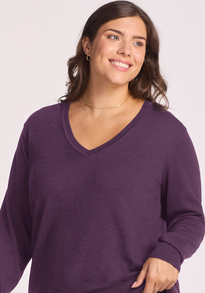 Woman with wavy brown hair smiles in a Woolx Francesca Sweater - Purple Iris and gold necklace, light background.