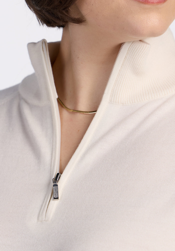 Close-up of a person wearing the Woolx Collins Half Zip Sweater in Dewdrop Pearl, collar partially unzipped to reveal a simple gold necklace. Only the lower face and upper chest are visible.