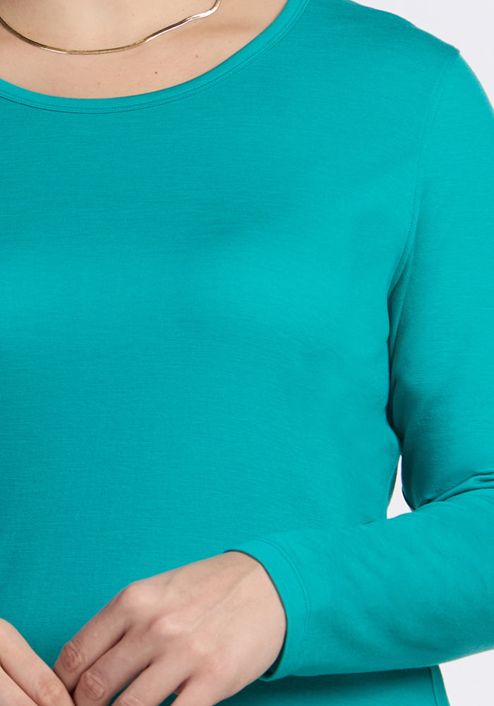 A person wearing the Woolx Remi Long Sleeve T-Shirt in Cool Jade and a thin gold necklace, with hands visible at the bottom of the image. The background is plain and light colored.