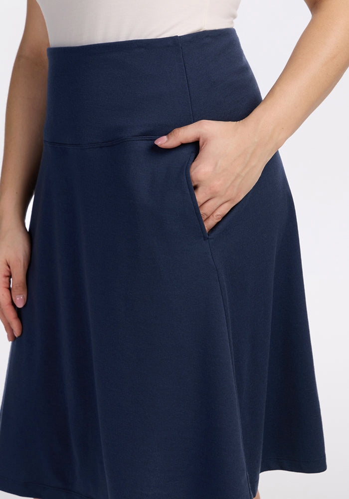 Wearing the Woolx Marilyn Flowy Midi Skirt in Deep Navy, one hand is placed in a pocket while the other rests by their side. The skirt, paired with a light top, combines stylish design and comfort.