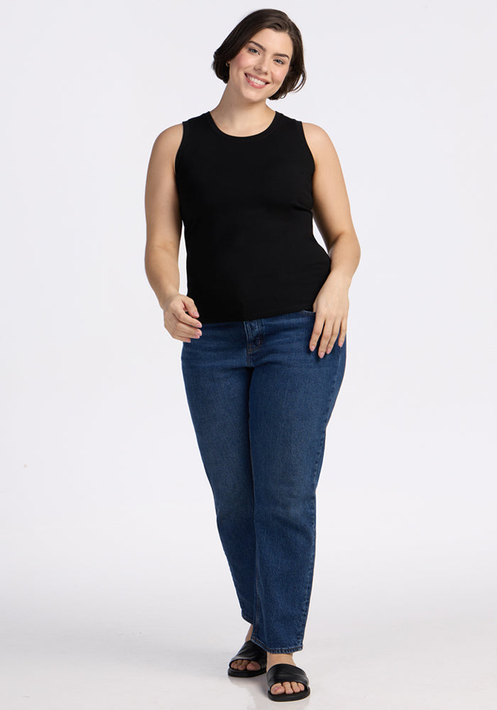 A woman with short brown hair smiles while standing against a plain white background. She wears the Woolx Ava Tank Top in black, blue jeans, and black sandals.