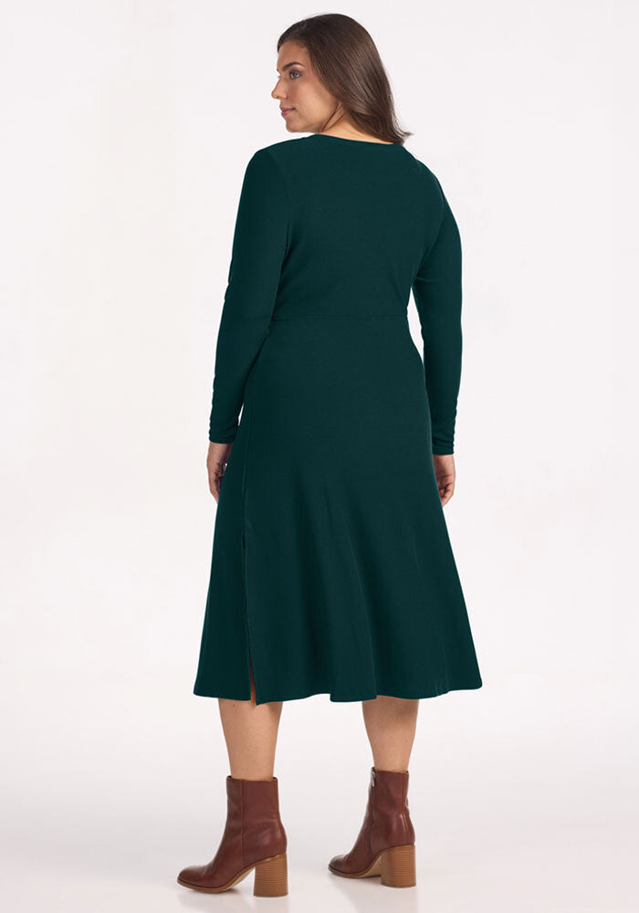 Woman in a Woolx Caroline Ribbed Twirl Dress in Deep Pine and brown heeled boots, standing with her back to camera.
