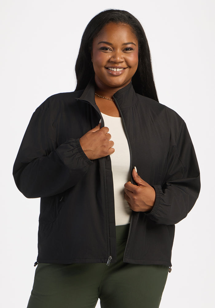 Woman smiling in Woolx Cooper Windbreaker - Black over a white top and green pants, against a plain background. 