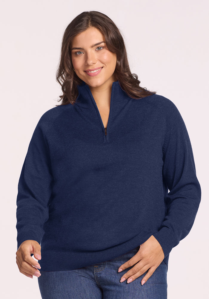 Smiling woman in Woolx Collins Half Zip Sweater - Ocean Cavern and jeans, one hand raised, against a light background. 