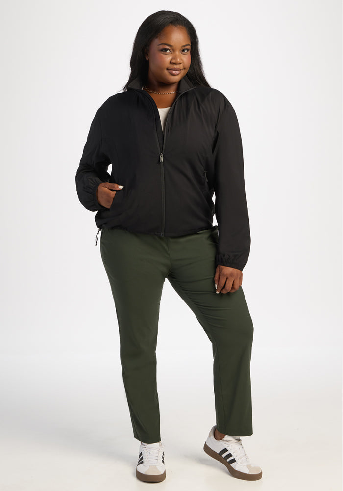 Woman wears Woolx Cooper Windbreaker in black, paired with green pants and white sneakers, hand in pocket.