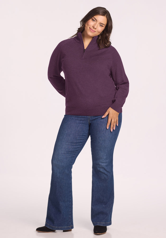 Woman in Woolx Collins Half Zip Sweater - Purple Iris and blue jeans stands, smiling with hand behind her back.