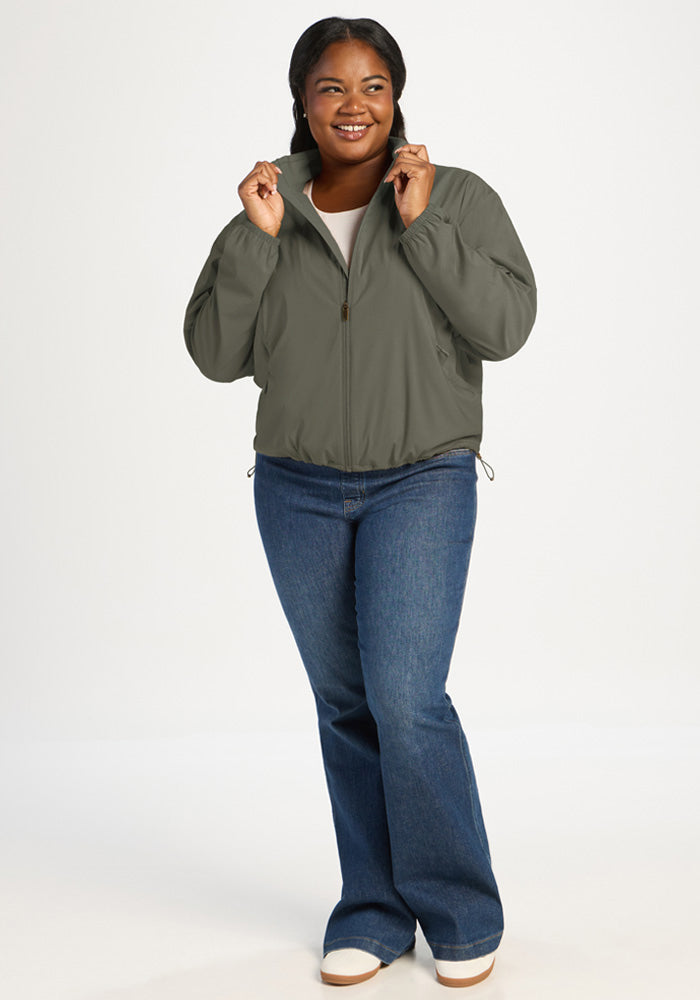A smiling woman wears an olive green Cooper Windbreaker - Sage Olive and blue jeans.